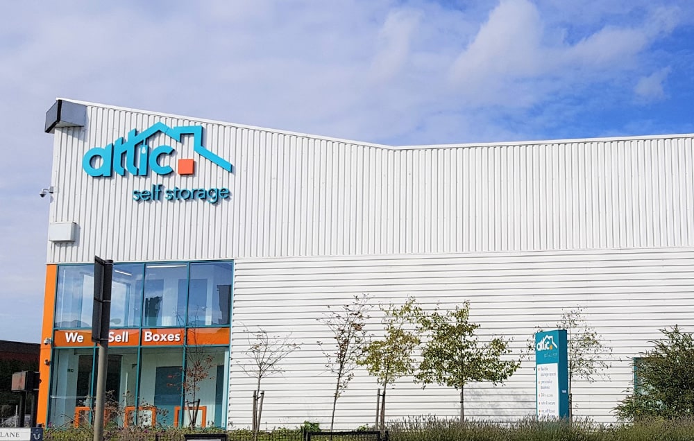 A large white building in Bow with a blue sign that says "Attic Self Storage" with trees in front of the building.